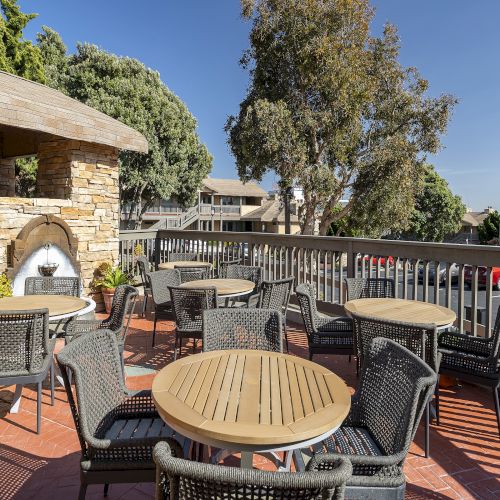 A sunny outdoor patio with wicker chairs and round wooden tables, a stone oven/barbecue, and a view of trees and buildings on a deck.