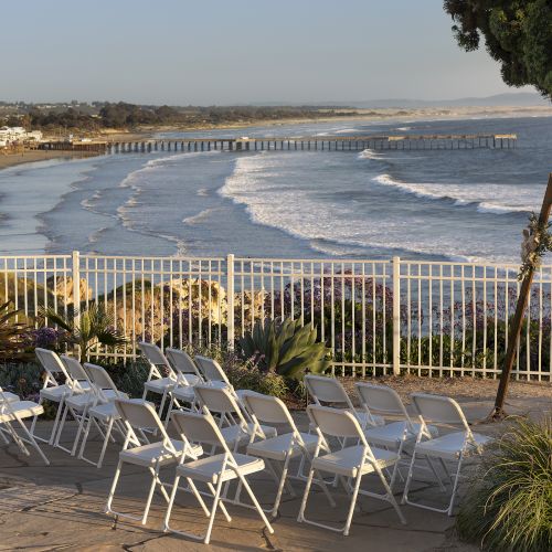 Beachside seating facing ocean waves, a fenced promenade, and a pier in the distance under clear skies.