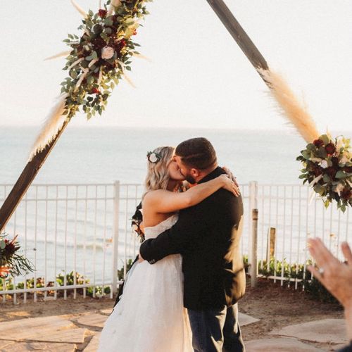 A newlywed couple shares a kiss under a triangular floral arch by the beach.