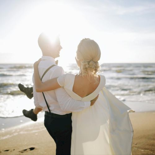 A couple in wedding attire hugs on a sunny beach, the ocean waves behind them, bright light haloing the scene as they stand close by the shore.