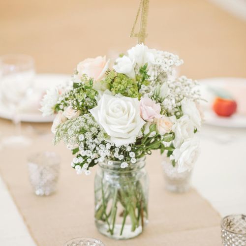 A centerpiece bouquet of white and pale pink roses with baby's breath in a mason jar, surrounded by votive candles on a beige tablecloth.