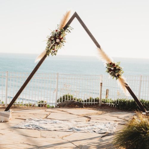 A wooden triangle arch decorated with flowers sits on a seaside terrace, a white cloth on the ground and ocean waves in the background.