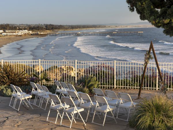 Chairs set up on a seaside patio overlook a wavy beach and pier in the distance; calm ocean, clear sky, and a tree in the foreground.