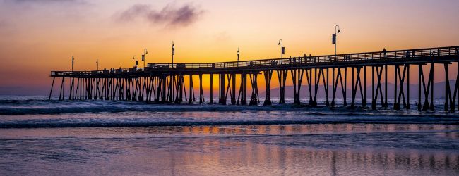 A seaside pier extending into calm water at sunset, with warm orange and purple skies reflecting on the wet sand and gentle waves.