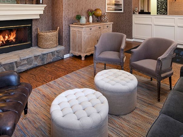 Cozy living room with a lit fireplace, stone hearth, leather chair, two gray armchairs, and two tufted round ottomans on a wood floor. End.