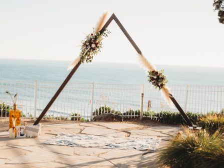 A minimal triangular wedding arch decorated with flowers by the seaside, on a stone terrace with a view of the ocean and a fence in the background.