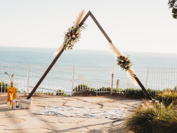 A minimal triangular wedding arch decorated with flowers by the seaside, on a stone terrace with a view of the ocean and a fence in the background.