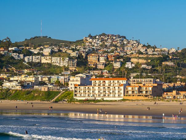 A coastal town on a hillside with pastel buildings, a sandy beach, and gentle waves under a clear blue sky.