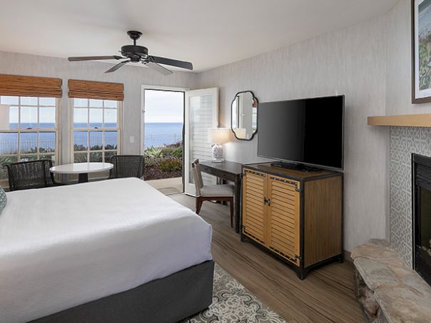 A bright hotel room with a cozy bed, fireplace, large windows overlooking the ocean, a TV on a wooden stand, and a small seating area.