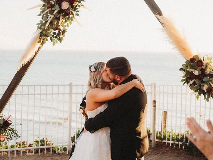 A couple shares a kiss at their beach wedding under a floral triangular arch, with guests nearby celebrating.